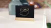 A small black chalkboard with a chalk drawing of a coffee cup on a wooden stand, placed on a white surface. Anteprima dell’immagine prodotto