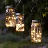 Three mason jar lanterns with fairy lights hanging from a branch outdoors at dusk. Produktbild-Vorschau