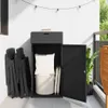 A black poly rattan garden storage cabinet is shown on a balcony, with its door open, revealing cushions and a basket inside. Folded chairs are stacked to the left of the cabinet. Náhľad obrázku produktu