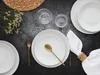 A table setting with white plates, gold cutlery, a white napkin, and a glass of water on a dark textured surface. Produktbild-Vorschau