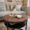 A round coffee table with a wooden top and black metal base, styled with a vase and faux plants, a candle holder with candles, and an open magazine. A light blue couch is visible in the background. Produktbild-Vorschau