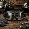 Paar Vintage-Leder-Motorradbrillen mit klaren Gläsern, auf rustikaler Holzoberfläche platziert, umgeben von anderen Anteprima dell’immagine prodotto