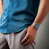 A person wearing a blue shirt and gray pants, with a silver watch on their wrist. Anteprima dell’immagine prodotto