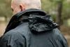 Close-up of the back of a person wearing a black jacket, with a hood folded up at the neck. Produktbild-Vorschau