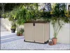 A beige and brown outdoor storage box is placed on a cobblestone patio next to a wall with lush green plants and ferns growing above it. Stairs are visible on the left side of the image. Voir les images du produit
