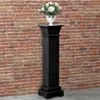 A black column stand with a vase of white flowers on top, set against a brick wall. Voir les images du produit