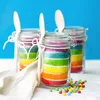 Three rainbow cakes in jars with wooden spoons and colorful candies. Náhled obrázku produktu