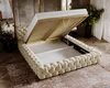 A tufted bed frame with its storage compartment open, revealing the slatted base and storage space. The bed is placed on a patterned rug in a room setting. Náhled obrázku produktu