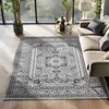 A living room scene with a large oriental rug as the focal point, a sofa, a coffee table with decorative items, and a large window with greenery visible outside. Produktbild-Vorschau