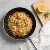 A close-up overhead view of a pan of spaghetti with shrimp and tomatoes, with a lemon and parsley on a cutting board to the side. Produktbild-Vorschau
