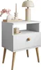A white nightstand with wooden legs, decorated with a lamp, a candle, and a vase with flowers. Produktbild-Vorschau