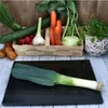 A black cutting board with a leek in the foreground, and a basket of vegetables in the background. Náhled obrázku produktu