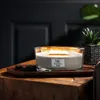 A lit WoodWick candle sits on a wooden table next to a potted plant. Podgląd zdjęcia produktu