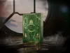A green and gold deck of Wicked playing cards sits on a dark, wet surface, with a large, circular window and smoke in the background. Náhled obrázku produktu