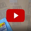 A collection of clear plastic templates for camera lens protectors, with a YouTube play button overlay and a cleaning wipe packet in the foreground. Produktbild-Vorschau