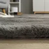 A close-up view of a gray shaggy rug on a wooden floor, with furniture and shelves blurred in the background. Anteprima dell’immagine prodotto