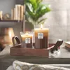 Two lit candles in glass holders sit on a wooden tray, with a blurred background of books and a plant. Náhled obrázku produktu