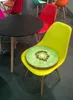 A yellow chair with a kiwi-shaped cushion in a room with other colorful chairs and a table. Produktbild-Vorschau