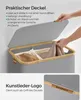 A person's hands are shown opening the lid of a grey laundry basket with a bamboo rim. The basket contains two compartments with laundry inside. A close-up of a leather logo on the lid is also shown. Produktbild-Vorschau