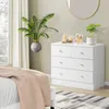 A white dresser with gold knobs sits next to a bed with a pink throw blanket. A large potted plant is on the left side of the dresser, and a small potted plant sits on top of the dresser. A round gold mirror hangs on the wall above the dresser. Produktbild-Vorschau