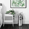 A fire extinguisher is placed on a wooden floor next to a white step stool with a plant on it, in front of a white wall with a framed picture. Produktbild-Vorschau