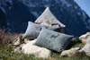 Three decorative pillows with deer motifs are arranged on rocks and a sheepskin rug, with a mountain landscape in the background. Produktbild-Vorschau