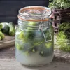 A Kilner preserving jar filled with pickles and dill, sitting on a wooden surface with some raw pickles and dill sprigs nearby. Anteprima dell’immagine prodotto
