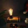 A vintage-style desk lamp with a wooden base and exposed filament bulb, placed on a dark surface with some decorative items in the foreground and background. Produktbild-Vorschau