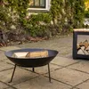 A black metal fire pit with logs inside, placed on a paved patio next to a wooden side table with more logs and two wine glasses. Produktbild-Vorschau