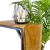 A close-up of a wooden side table with a black metal frame, featuring a geometric black lantern with a lit white candle on top. Green foliage is visible in the background. Voir les images du produit