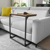 A C-shaped side table with a wooden top is placed next to a light gray sectional sofa in a living room. Produktbild-Vorschau