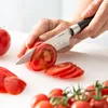 A person is slicing a tomato on a white cutting board with a knife. Produktbild-Vorschau