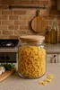 A glass jar filled with farfalle pasta sits on a kitchen counter, with a wooden cutting board and garlic in the foreground, and kitchen appliances and brick wall in the background. Produktbild-Vorschau