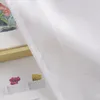 A close-up view of a white sheer curtain with a subtle textured pattern. In the background, a framed picture with floral elements and small potted plants is partially visible. Produktbild-Vorschau