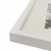 A close-up, angled view of a white picture frame with a white matting, showing a portion of a black and white photograph within. Náhľad obrázku produktu