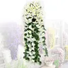 Weiße künstliche Hängeblumen mit grünen Blättern, präsentiert vor einem unscharfen Hintergrund aus weißer Ziegelwand und anderen bunten Kunstpflanzen Náhled obrázku produktu