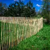 A rustic wooden fence made of vertical stakes, set in a grassy field with trees and a blue sky in the background. Produktbild-Vorschau