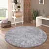 A round, gray, shaggy rug is placed on a wooden floor in a living room setting. A ladder shelf with decorative items, a basket with a pillow, and a plant are visible in the background. Anteprima dell’immagine prodotto