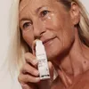 A woman is holding a small bottle of eye cream and applying it to her under-eye area. Produktbild-Vorschau