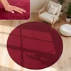 A round, red rug is placed on a wooden floor in a living room setting, with a chair and plants in the background. Produktbild-Vorschau
