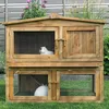 A two-story wooden rabbit hutch with two rabbits inside, set on a grassy lawn with a fence and plants in the background. Produktbild-Vorschau