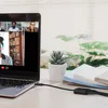 A laptop displaying a video conference call, with a USB-C adapter and cable connected to it, placed on a desk with a small plant and books. Produktbild-Vorschau