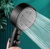 A hand holding a black shower head with water spraying out, with large green leaves in the background. Produktbild-Vorschau