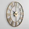 A close-up, angled view of a large, round, gold-colored metal wall clock with Roman numerals, mounted on a light gray wall. Produktbild-Vorschau