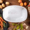 A white ceramic roasting dish with the word Römertopf embossed on it, surrounded by ingredients like potatoes, carrots, and herbs on a wooden table. Produktbild-Vorschau