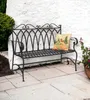 A metal garden bench with a decorative pillow sits on a stone patio next to a white wall and greenery. Anteprima dell’immagine prodotto