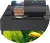 An automatic fish feeder sits on top of a black aquarium lid, with a yellow fish swimming in the aquarium below. Produktbild-Vorschau