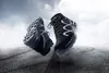 Two black safety boots are shown in mid-air above a wet asphalt road with tire tracks, under a cloudy sky. Produktbild-Vorschau