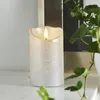 A close-up of a silver LED candle with a flickering flame, placed on a white surface with a plant in the foreground and a blurred background. Podgląd zdjęcia produktu