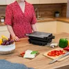 A person in a red dress is standing in a kitchen next to a panini grill, with ingredients for making sandwiches. Produktbild-Vorschau